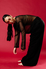 Asian woman in black pants and sheer top bending over on a red background with a wall behind her