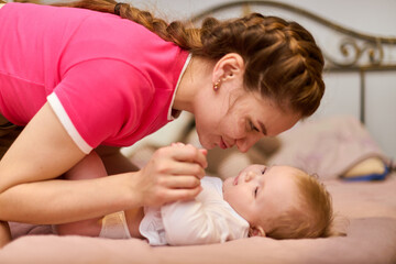 Young woman with braided hair and baby girl interact warmly on soft pink bed. Woman in pink top kisses baby girl, creating tender bond. Warm indoor light enhances comfort