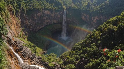 Aerial video captures a stunning waterfall cascading into a lush, green canyon, with vibrant rainbows forming in the mist, viewed from above.