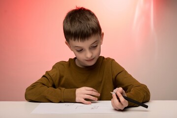 Naklejka premium Young boy in brown sweater drawing at a desk, pink background. Ideal for themes of childhood creativity, education, focus, and artistic expression.