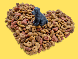 Black toy cocker spaniel sitting on a heart shaped pile of dry dog food on yellow background