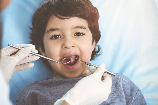Cute arab boy sitting at dental chair with open mouth during oral checking up with doctor. Visiting sunny dentist office