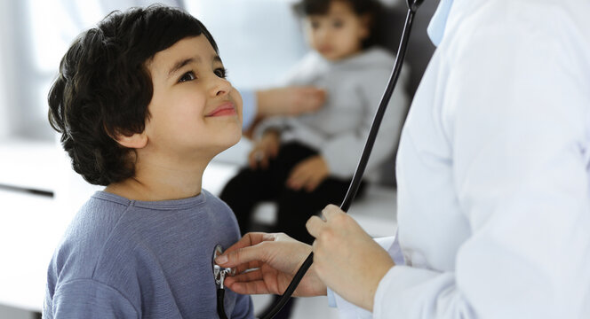 Doctor-woman examining a child patient by stethoscope. Cute arab boy and his brother at physician appointment. Medicine concept
