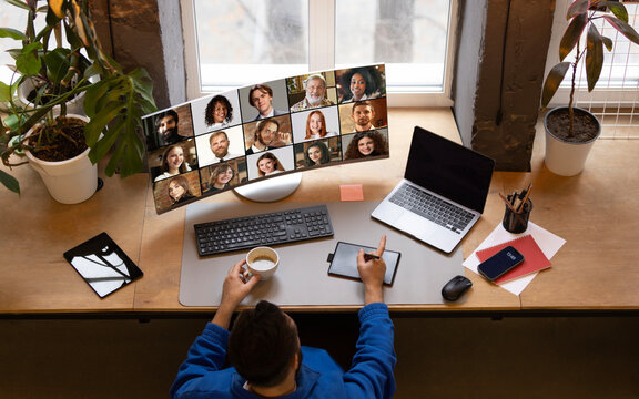 Overhead view of remote worker in blue jacket holding coffee, engaging in online meeting with large virtual team displayed on monitor against cozy home office. Concept of teleworking, networking.