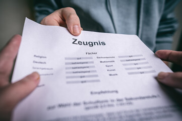 Nahaufnahme einer Zeugnisübergabe. Eine Person reicht einer anderen ein Schulzeugnis mit Notenübersicht. Das Bild steht für Bildung, Schulabschluss, Leistungsbewertung und akademische Erfolge.