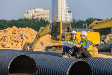 Two engineer sitting on large pipes at a construction site, reviewing blueprints. A large excavator is visible in the background, and the construction area is set in an urban environment.