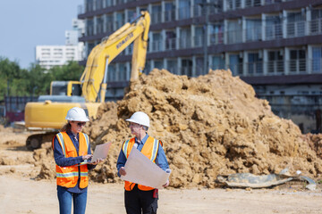 supervisor standing at a construction site, reviewing blueprints. A large pile of dirt and an...