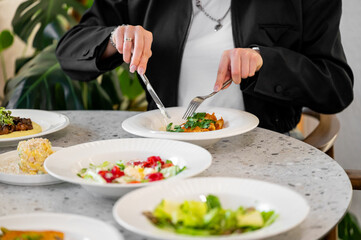 A woman elegantly dining at a table, using a knife and fork to savor a delicious meal. Fresh salads and dishes surround her, highlighting a beautiful dining experience