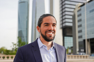 Fototapeta premium Business man smiling in front of camera with financial skyscraper in background - Confident person in the city - Focus on face