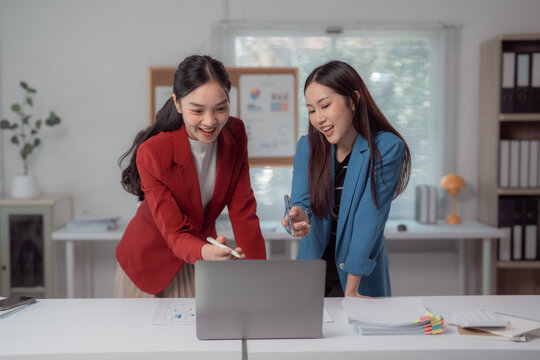 Two happy asian businesswomen discussing and working together using laptop computer while standing at desk in modern office, entrepreneurship and collaboration concept