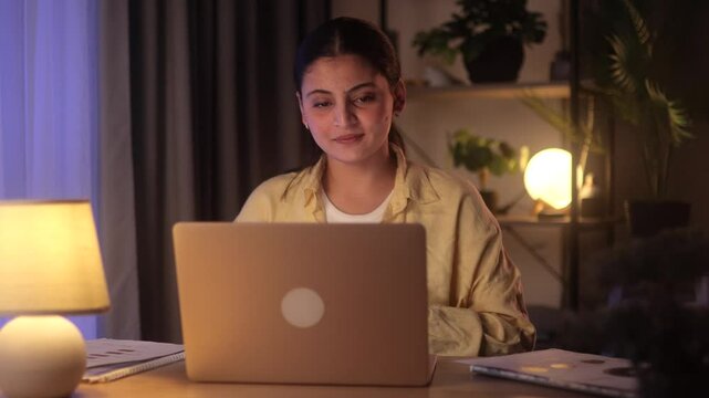 Smiling Indian woman having a friendly video call on her laptop at night. Engaging in an online conversation in a cozy home office setting