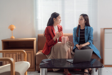 Two asian businesswomen are discussing a project, sitting on a sofa in a modern office, with a...