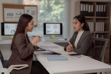 Two asian businesswomen are discussing marketing strategy using data charts and graphs, collaborating on a project in a modern office, showcasing teamwork and data analysis