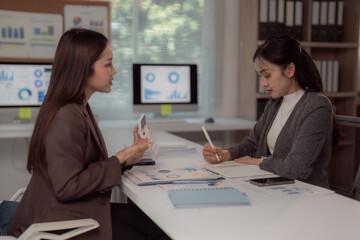 Two young businesswomen are sitting at office desk discussing and analyzing data using calculator and taking notes of financial graphs and charts displayed on computer screen