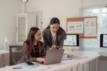 Two businesswomen work together in a modern office, focusing on a laptop and discussing data. Charts and graphs are visible on screens and a bulletin board, indicating a data-driven environment