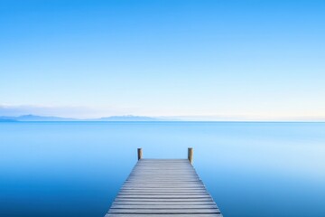 Fototapeta premium Wooden dock extends into calm, blue waters under a clear sky, mountains visible on the horizon