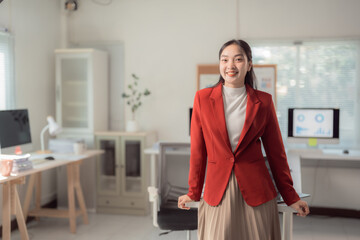 Confident young woman in a red blazer leaning against a desk in a bright, modern office. The...