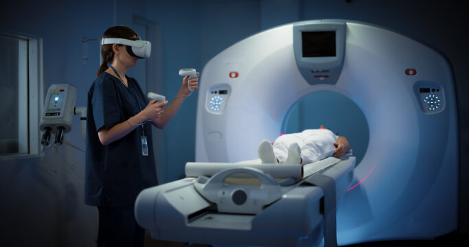 Futuristic Concept: In Medical Laboratory Room, Female Doctor Wearing Virtual Reality Headset Monitors Patient Undergoing MRI or CT Scan Procedure. Healthcare Facility with High Tech Equipment