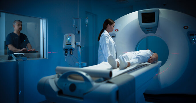 High-Tech Modern Medical Equipment: In Medical Laboratory Female Radiologist and Male Doctor Control and Monitor MRI or CT Scan with Female Patient Undergoing Procedure