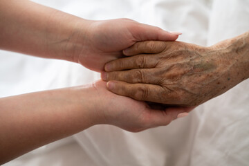 A young womans hands gently hold the hands of an elderly female patient in a hospital bed,...