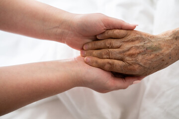 A young womans hands gently hold the hands of an elderly female patient in a hospital bed, symbolizing care, support, and compassion.