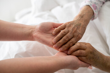A young womans hands gently hold the hands of an elderly female patient in a hospital bed, symbolizing care, support, and compassion.