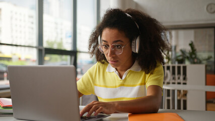 African American woman student girl ethnic female wearing headphones writing homework prepare exams test studying online video class lesson internet with laptop indoors school university e-learning