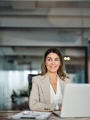 Happy mid aged professional business woman working in office with laptop looking away at copy...