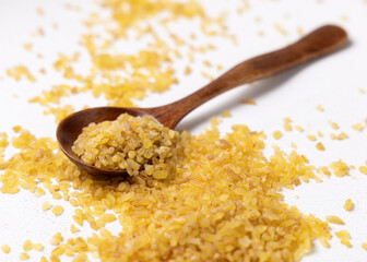 Raw dry uncooked yellow bulgur grain in a wooden spoon on white table cloesup. Ancient food