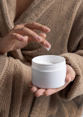 Woman in brown bathrobe touching cream with finger and holding a cosmetic jar closeup
