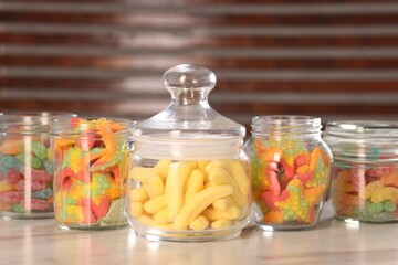 Tasty gummy candies in glass jars on white marble table, closeup