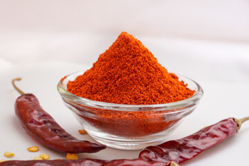 Chili powder is a seasoning blend of ground dried chills in a glass bowl with dried red chills on a white background.