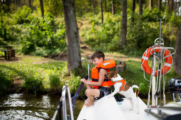 Młody chłopiec ubrany w pomarańczową kamizelkę asekuracyjną śmieje się i bawi na jachcie z rodzicami, bezpieczne wakacje na wodzie i jachcie © Katarzyna Krociel