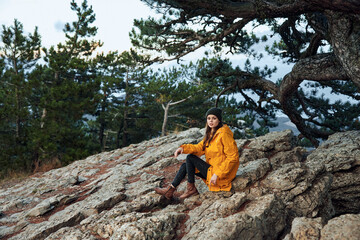 Woman in a vibrant yellow coat enjoying nature's beauty while sitting on a rock near a majestic pine tree