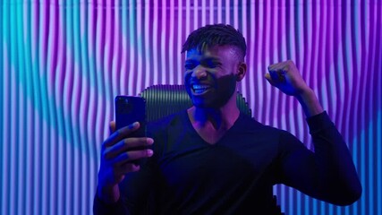 African American man guy male indoors futuristic innovation cyberspace neon lights winning with mobile phone bet casino online crypto win victory champion celebrate yelling happy video game addict