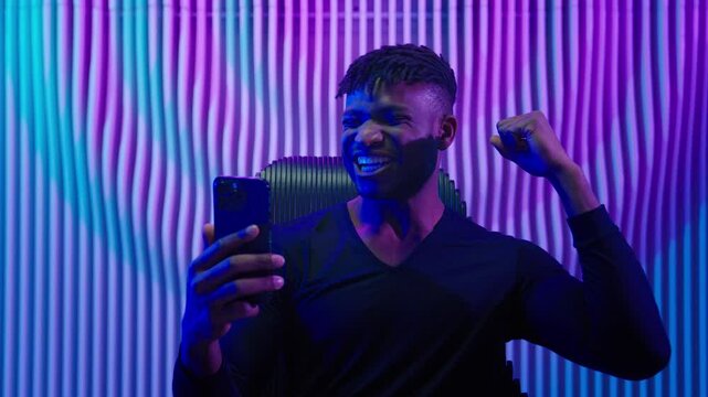 African American man guy male indoors futuristic innovation cyberspace neon lights winning with mobile phone bet casino online crypto win victory champion celebrate yelling happy video game addict