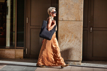 Stylish Mature Woman with Short Hairstyle Walks, Wearing Summer Beige maxi Dress and Large black...