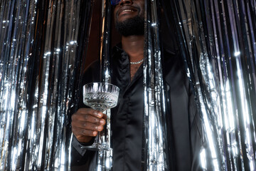 Person standing behind shimmering curtains while holding a crystal glass in hand, partially hidden...