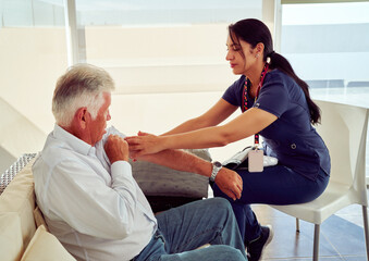 Young nurse examining senior man's arm during home visit, providing medical assistance and...
