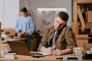 Man sitting at desk in office, using phone and laptop for work, shelves with boxes and woman in background, creating busy and productive atmosphere