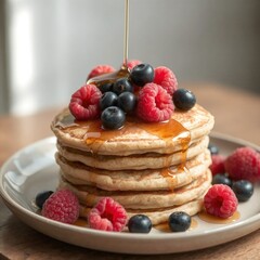 A stack of fluffy oat pancakes topped with fresh raspberries, blueberries, and a drizzle of maple syrup, served on a neutral ceramic plate. 