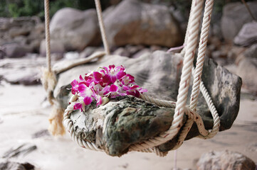 A rustic wooden swing with purple flowers on a tropical beach, surrounded by rocks and trees.