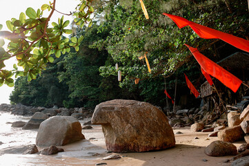 A serene beach scene with large rocks, lush greenery, and red flags hanging from trees. The sunlight filters through the leaves, casting gentle shadows on the sandy shore.