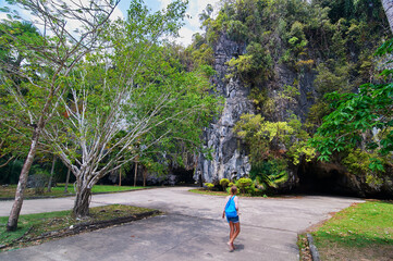 A person walks towards a large limestone cave entrance surrounded by lush greenery.