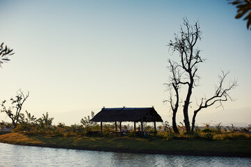 Fototapeta premium A serene lakeside view with a small island featuring trees and a wooden gazebo, surrounded by calm water and distant buildings under a clear blue sky.