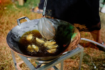 A person cooking traditional street food in a wok at a market, using a spatula to flip a crepe-like dish. Various ingredients and cooking utensils are visible around the cooking area.