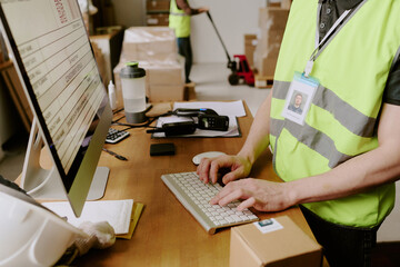 Worker wearing safety vest typing on keyboard in warehouse office while using various devices and...