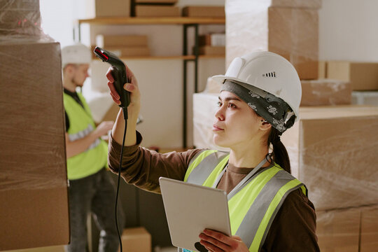 Woman in construction gear scanning bullet barcode with handheld scanner while holding tablet in warehouse environment filled with packed boxes and shelves - Powered by Adobe