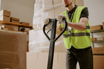 Worker in high-visibility vest operating pallet jack in warehouse, surrounded by stacked cardboard boxes and shelves, performing shipping and handling tasks for logistics operations