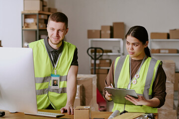 Two workers wearing safety vests in warehouse setting, engaging in collaborative tasks, shelves lined with boxes and equipment in background, demonstrating efficient teamwork